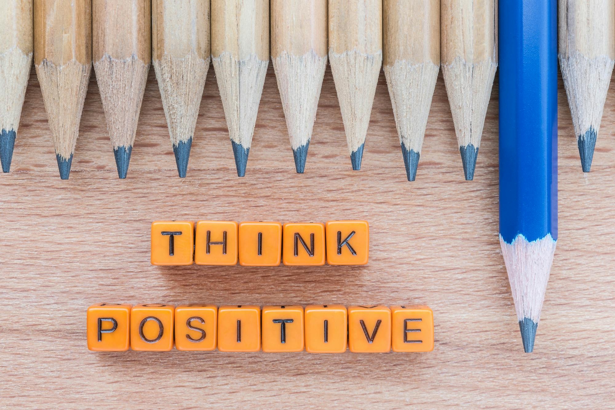 Imagen donde se ve una fila de lápices marrones sobre una mesa, excepto uno de color azul que sobresale de las demás, y debajo hay un texto que dice "think positive", que se traduce a "piensa positivo".