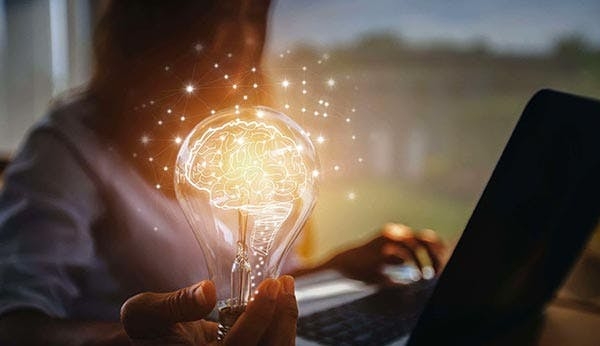 Imagen de una mujer, trabajando en su laptop, mientras sostiene una bombilla iluminada que representa la creatividad e innovación.